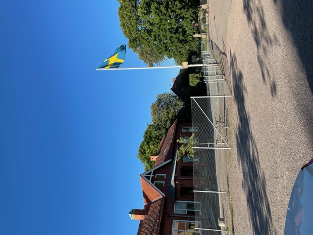 Bild av en hissad svensk flagga på en skolgård i sommarväder.